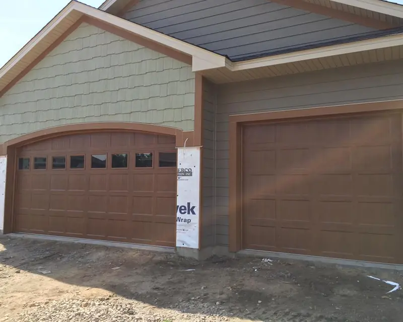 Brown carriage-style double and single garage doors on a newly built home still wrapped in house wrap