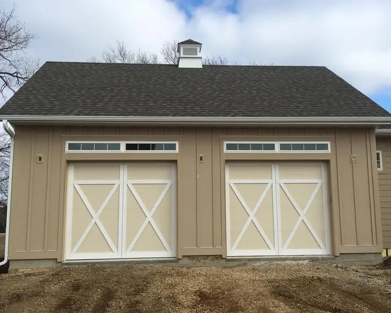 Two tan carriage-style garage doors with white crossbuck trim and top windows on a detached garage
